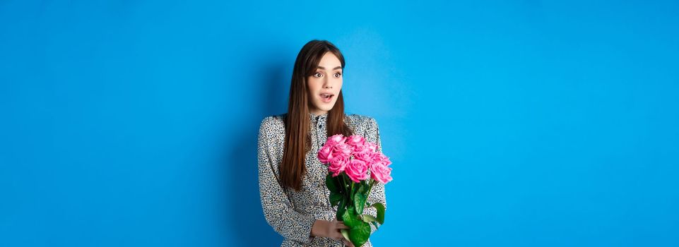 Valentines day concept. Surprised happy girl looking aside with amazement, receiving bouquet of romantic flowers, holding pink roses, standing in dress on blue background.