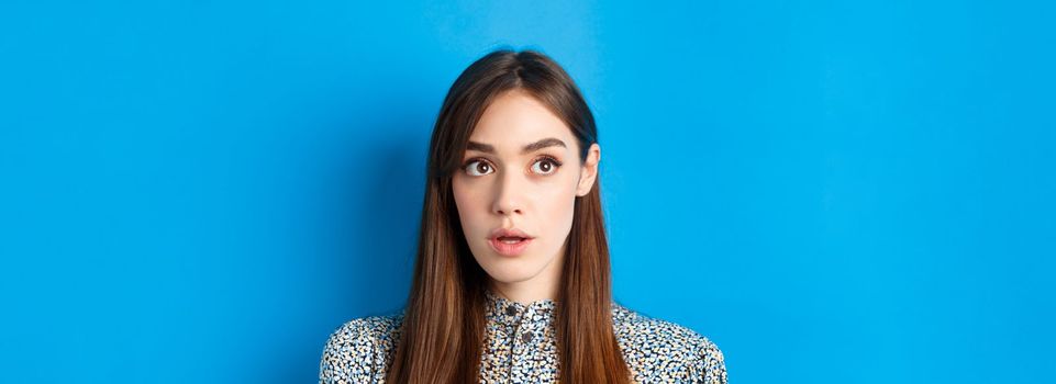 Close-up portrait of surprised young woman with natural beauty, looking at upper left corner and gasping, standing on blue background.