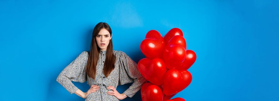 Valentines day. Angry and confused girlfriend in dress, standing near red heart balloons and frowning annoyed at camera, standing near blue background.