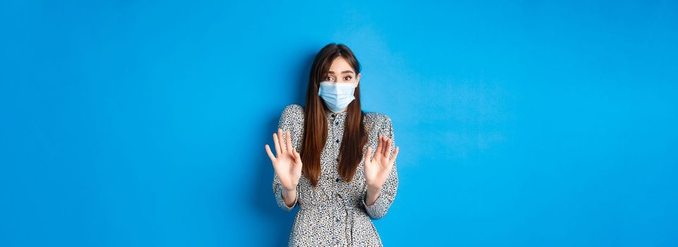 Covid-19, pandemic lifestyle concept. No thank you. Reluctant and worried girl in medical mask asking non-masker to stay away, showing block gesture, tell stop, blue background.