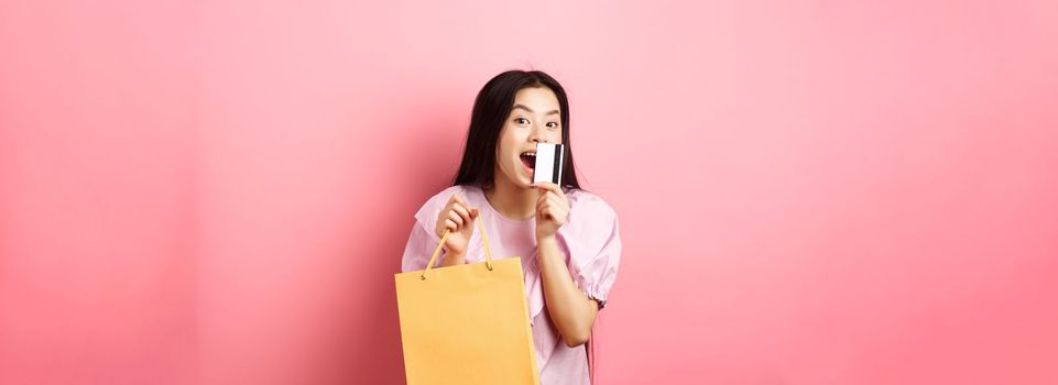 Shopping. Excited beautiful asian woman buying in stores, holding paper bag and kissing plastic credit card, standing against pink background.