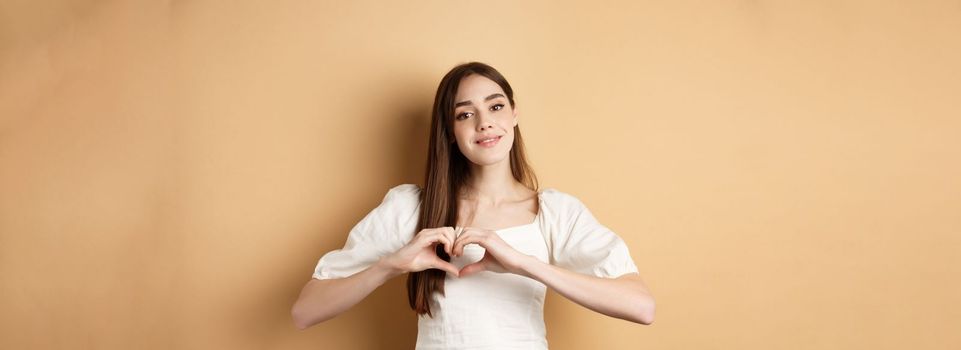 Beautiful caucasian woman say I love you, showing heart gesture and smile at camera, beige background. Concept of romance and Valentines day.