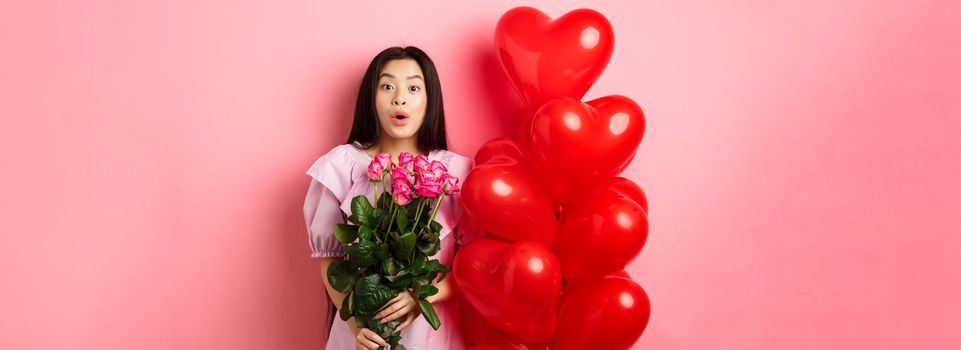 Surprised asian girl in dress standing near valentines day heart balloons and say wow at camera, holding flowers bouquet from lover, romantic date with roses, pink background.