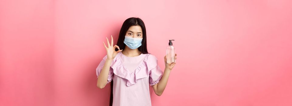 Healthy people and covid-19 pandemic concept. Cheerful asian woman in medical mask recommend hand sanitizer, showing okay sign, good antiseptic bottle, white background.
