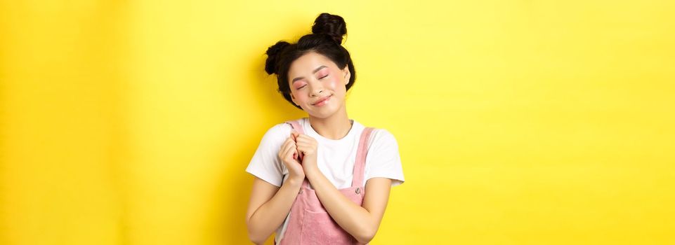 Cute asian girl with makeup, close eyes and remember beautiful moment, holding hands on heart daydreaming, standing on yellow background.