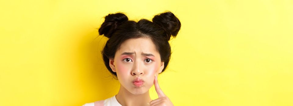 Close-up portrait of stylish asian girl with bright makeup and glowing skin, pouting and frowning, poking her cheek with sad face, standing upset on yellow background.