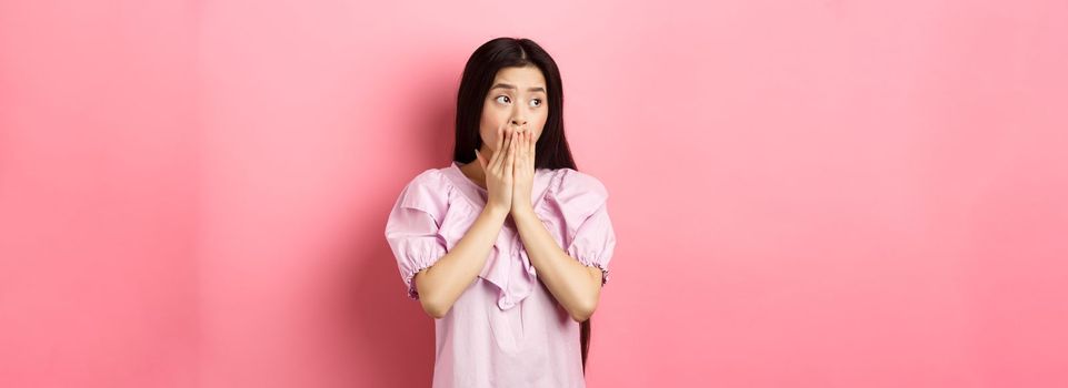 Shocked and worried asian woman looking aside at empty space with concerned face, covering mouth with hands, standing in dress on pink background.