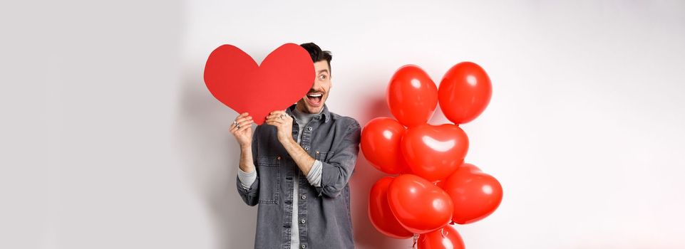 Romantic cheerful man cover half of face with Valentine heart cutout and smiling amazed, celebrating love holiday, standing on white background.