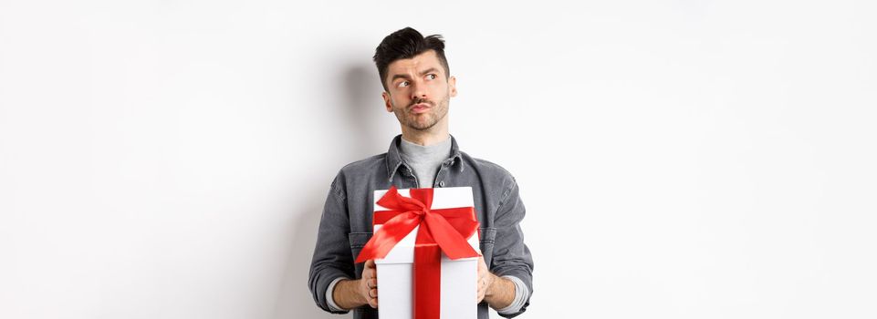 Pensive boyfriend looking aside and holding gift box, waiting for lover, making surprise present on valentines day, planning date with girlfriend, white background.