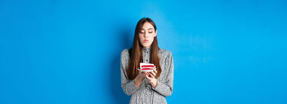 Cute birthday girl celebrating, blowing candle on cake and making wish, standing on blue background. Party and holidays concept.