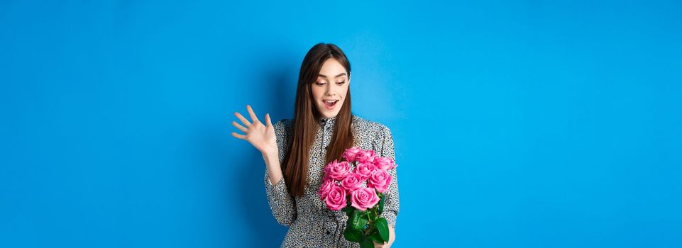 Valentines day concept. Image of attractive young woman gasping amazed, receive surprise flowers, standing on blue background.