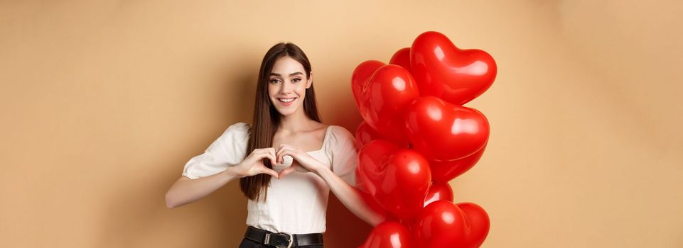 Lovely caucasian girl express love and tenderness, showing heart gesture and smiling, standing near Valentines day balloons, beige background.