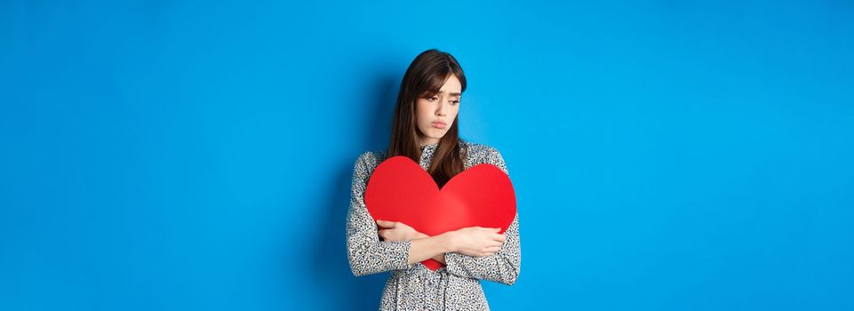 Valentines day. Lonely single girl hugging big red heart cutout and looking down upset, standing gloomy on blue background.