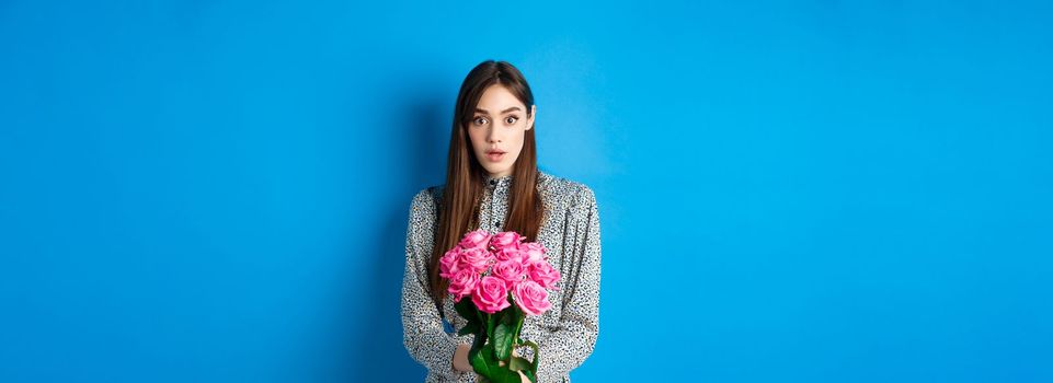 Valentines day concept. Surprised girlfriend receiving beautiful bouquet of flowers and looking with disbelief at camera, standing on blue background.