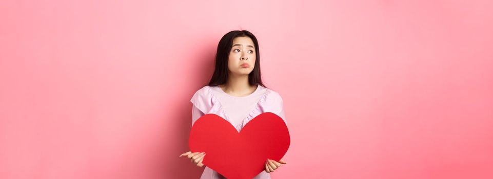 Valentines concept. Lonely teenage asian girl dreaming about love, feeling sad and lonely on lovers day, looking aside with pity, holding big red heart, pink background.