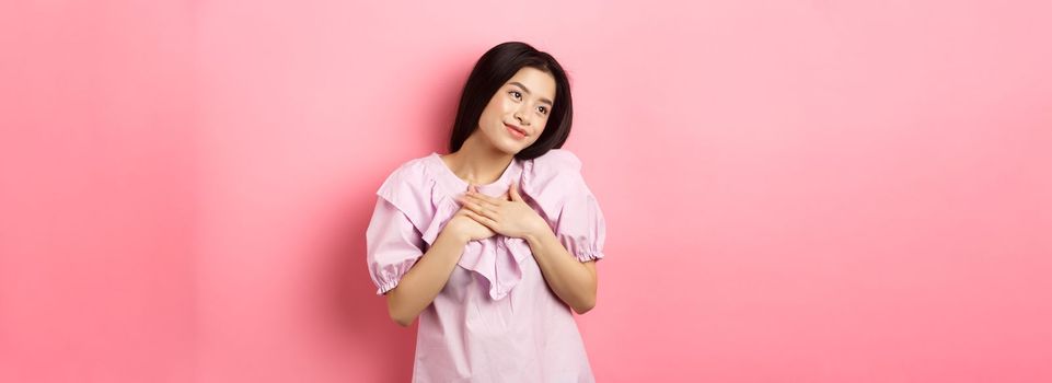 Dreamy asian girl smiling and holding hands on chest, looking left at something romantic and cute, standing on pink background.