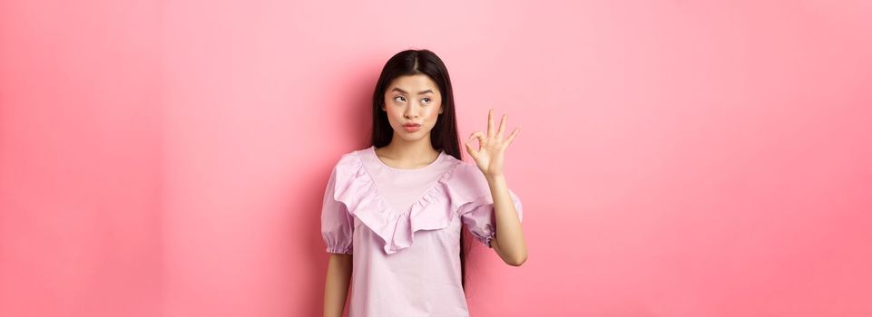 Sassy asian woman in dress show okay sign, looking aside, assure everything good, guarantee all OK, standing pleased on pink background.