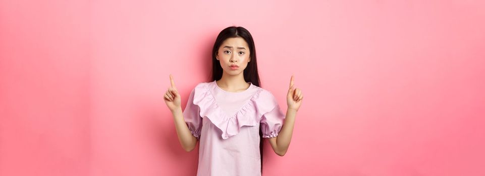 Cute timid asian woman pointing fingers up, frowning and look upset, pointing fingers up at logo, standing in dress on pink background.