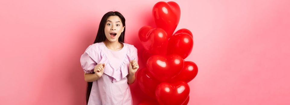 Excited and surprised teenage korean girl open mouth amazed, receive surprise gift on valentines day, looking wondered, standing near heart balloons, pink background.