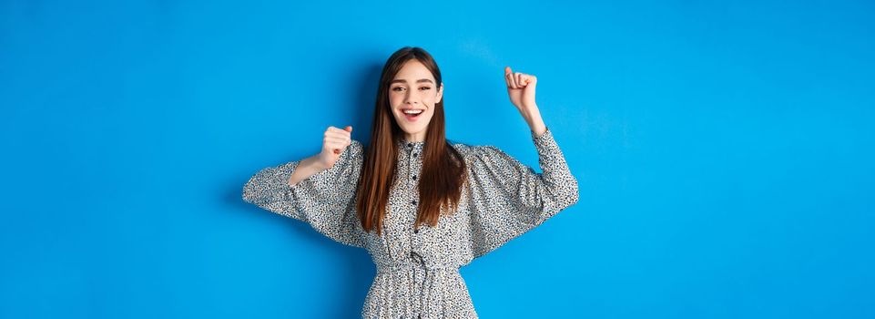 Cheerful young woman in dress raising hands up and smiling excited, triumphing over winning prize, celebrating victory, standing on blue background.