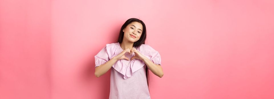 Valentines day concept. Beautiful korean teenager in dress showing heart gesture and smiling with admiration and affection, falling in love, feel romantic, pink background.