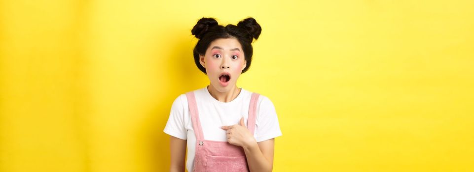Surprised asian teen girl with glamour makeup, pointing at herself and gasping confused, being chosen or accused, standing on pink background.