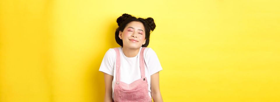Romantic asian girl with cute pink makeup and summer clothes, close eyes and smiling dreamy, standing on yellow background.