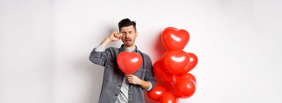 Valentines day and love concept. Sad crying man holding red heart balloon and whiping tears, standing single and miserable, being heartbroken, white background.