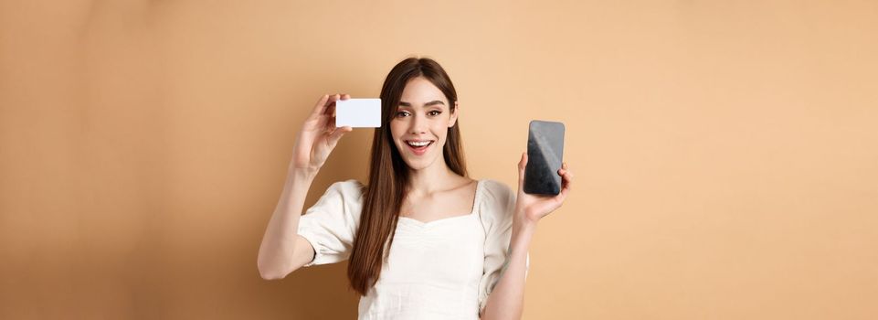 Cheerful young woman showing plastic credit card and empty mobile phone screen, smiling pleased at camera, standing on beige background.