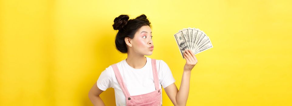 Shopping. Silly korean girl with glam makeup, pucker lips and looking at dollar bills, wasting money, standing on yellow background.