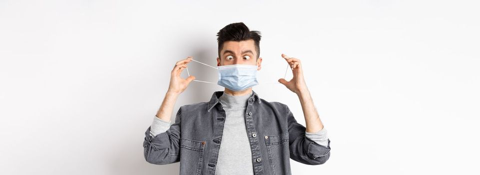 Health, covid and pandemic concept. Funny man squinting eyes at medical mask, playing with it, standing on white background.