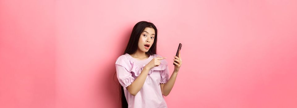 Excited beautiful asian woman, pointing finger at smartphone and look curious, checking out online promo, standing against pink background.