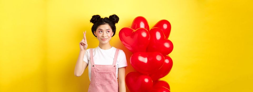 Valentines day concept. Beautiful korean girl dreaming of perfect date, making wish and looking at upper left corner, standing near red hearts balloons, yellow background.