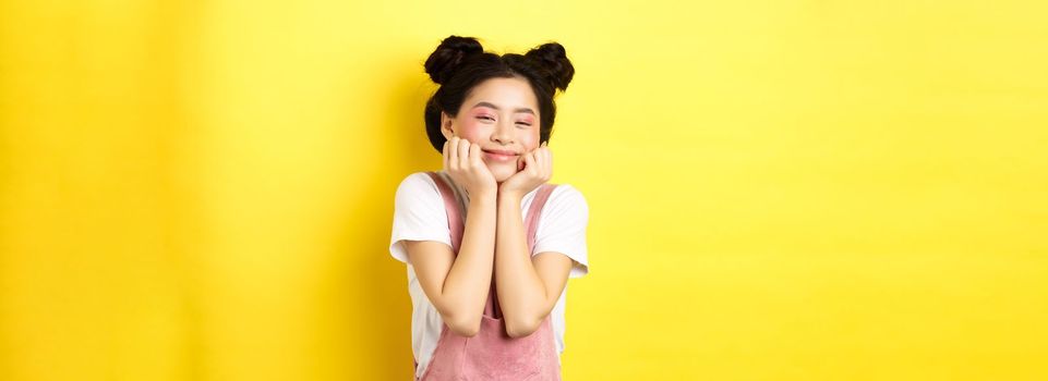 Cute happy asian woman with bright makeup and dungarees, leaning satisfied on hands and smiling, standing on yellow background.