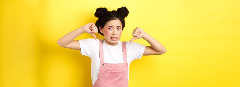 Disgusted asian girl showing thumbs down and cringe from bad product, standing in summer clothes against pink background.