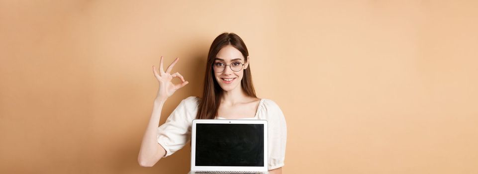 E-commerce. Smiling young woman in glasses showing okay sign and laptop screen, recommending internet promo, standing on beige background.