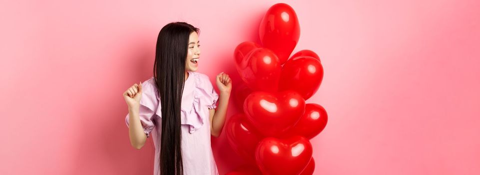Asian teenage girl with long hair, cheering from valentines day romantic gift, looking at logo and smiling happy, jumping from joy near lovers gift heart balloons, pink background.