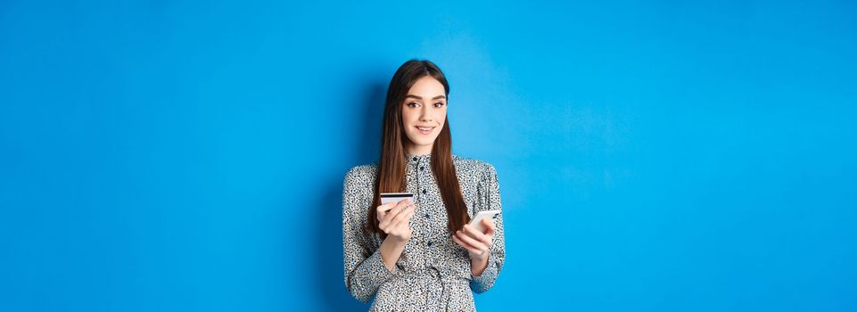 Online shopping. Happy smiling woman buying in app, holding smartphone and plastic credit card, blue background.
