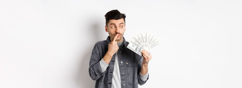 Man thinking of shopping while looking at dollar bills, holding money and staring pensive, standing on white background.