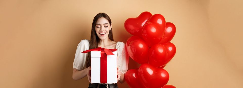 Happy woman open Valentines day gift, smiling excited and looking at present box, standing near red heart balloons on beige background.
