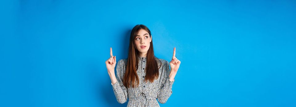 Pretty thoughtful woman pointing fingers up and thinking, standing pensive in dress on blue background.