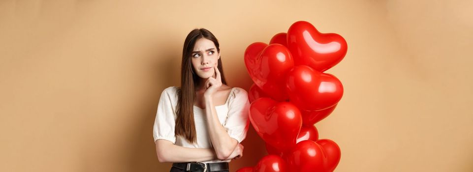 Valentines day and love concept. Pensive woman in trendy clothes and makeup, thinking and frowning, looking aside doubtful, standing near heart balloons, beige background.
