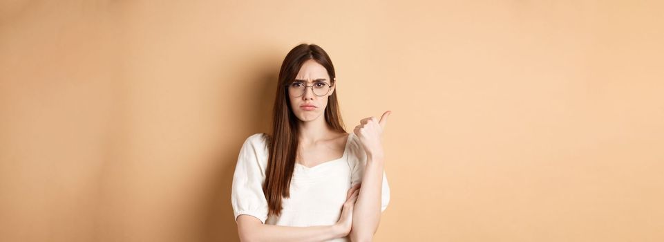 Disappointed girl in glasses frowning, pointing aside at bad product, disapprove and dislike something, standing on beige background.