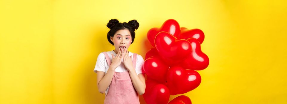 Surprised korean girl gasping and saying wow, looking at special promo offer on Valentines day, standing near big red hearts balloons and yellow background.