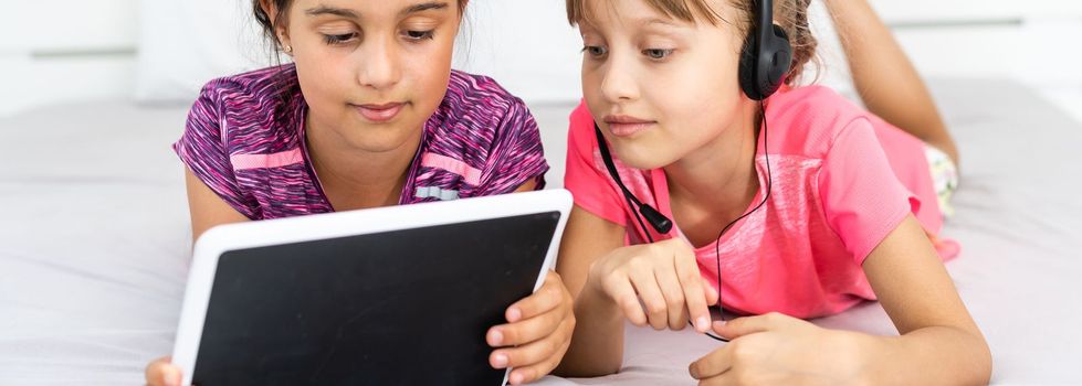 Two cute little girls are using a digital tablet and smiling while lying on bed in children's room.