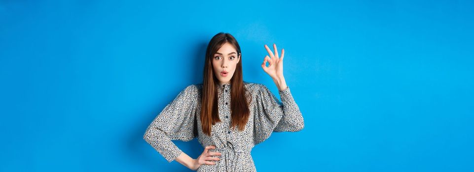 Impressed caucasian woman in dress showing okay sign, look with amazement and say wow, praise awesome product, standing on blue background.