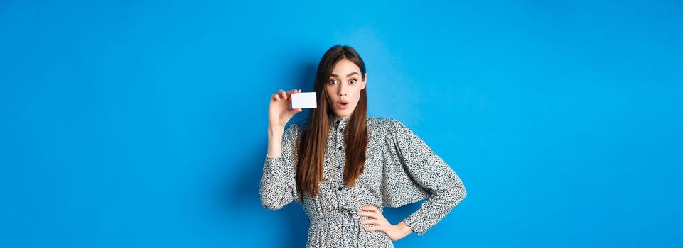 Shopping. Excited girl showing her plastic credit card and look at discounts, say wow amazed, standing on blue background.
