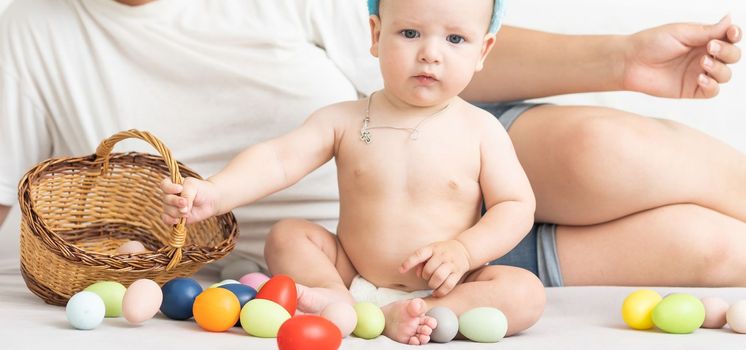 Mom and baby with rabbit ears, with Easter eggs in their hands, sitting on the sofa, parents and children play indoors. Family celebrates Easter.