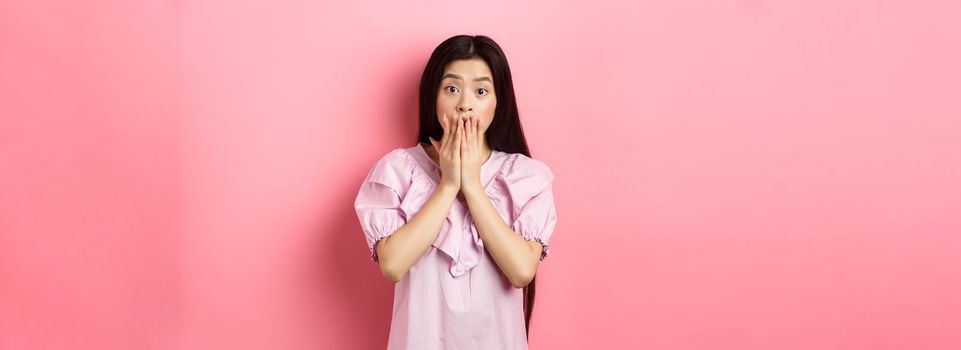 Shocked asian girl with long dark hair, gasping and covering mouth with hands, look at bad news, terrible accident, standing on pink background.