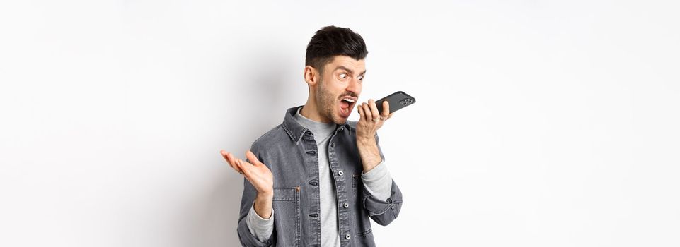 Angry guy shouting at speakerphone, scream on mobile phone, leaving angry voice message, standing against white background.